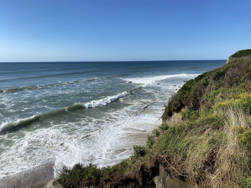 A scenic view of a coastline with waves crashing on the shore, surrounded by greenery under a clear blue sky.