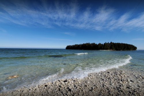 A serene shoreline with clear water, gentle waves, and a distant island surrounded by a blue sky.