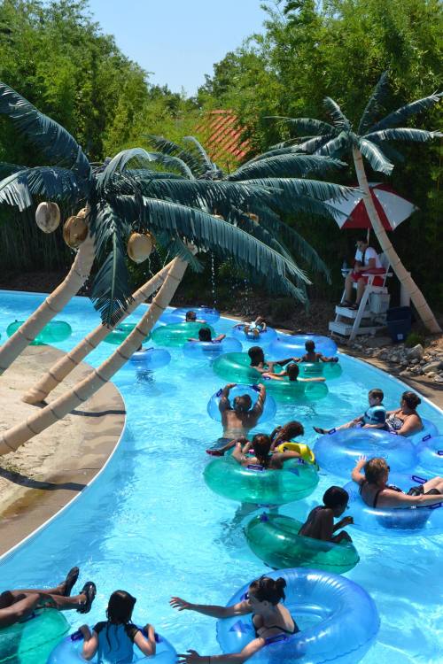 A water park scene with people floating on inner tubes in a winding pool surrounded by palm trees.