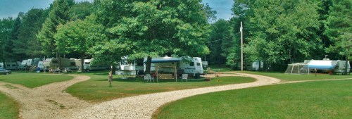A scenic campground with RVs parked along a gravel path, surrounded by lush green trees and open grass.