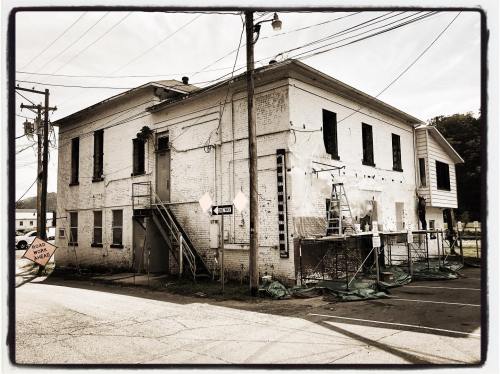 Abandoned building with peeling paint, broken windows, and construction equipment outside, surrounded by power lines.