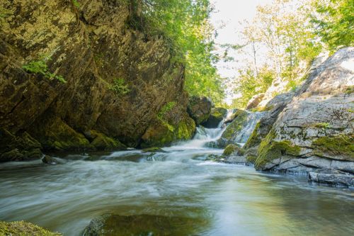 A serene river flows through rocky terrain, surrounded by lush greenery and gentle waterfalls under a bright sky.