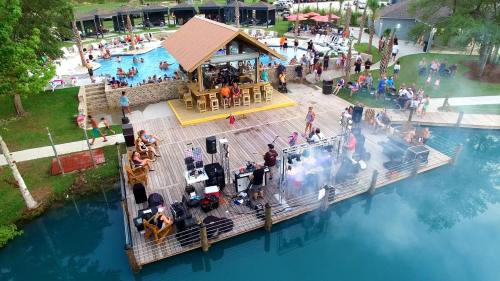 A lively outdoor scene with a band performing on a deck by a pool, surrounded by people enjoying the water and festivities.