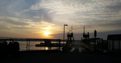 Sunrise over a calm waterfront with a ferry terminal and clouds in the sky, framed by a fence and light posts.