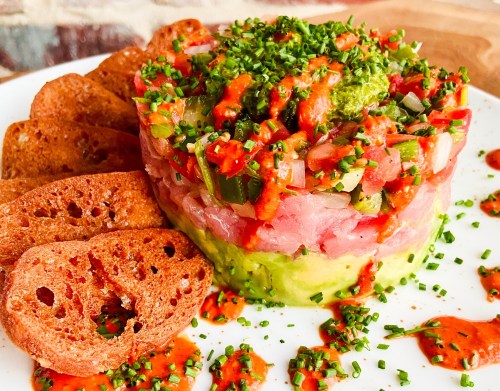 A colorful dish featuring layered tuna tartare with avocado, topped with herbs and sauce, served with crispy bread slices.