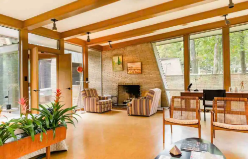 Modern living room with large windows, wooden beams, striped chairs, and a cozy fireplace surrounded by greenery.