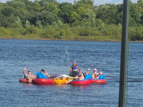 A group of people relax on colorful inner tubes in a lake, enjoying a sunny day and waving at the camera.