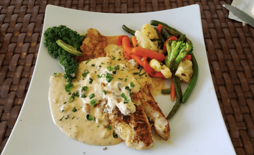 A plate of seasoned fish with creamy sauce, served with steamed vegetables and a side of broccoli.