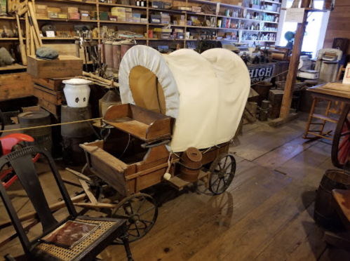 A vintage wooden wagon with a white canvas cover, displayed in a rustic museum filled with various historical items.