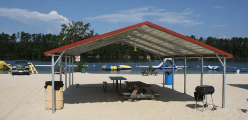 A shaded picnic area on a sandy beach, with a grill and picnic table, overlooking a lake and water activities.