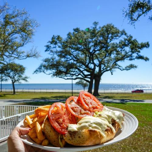 A hand holds a plate with a sandwich topped with tomatoes and fries, set against a sunny beach and tree backdrop.