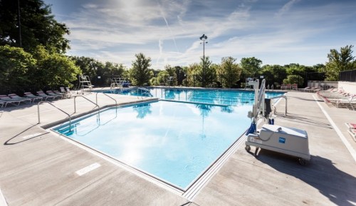 A clear swimming pool surrounded by lounge chairs and trees, under a bright blue sky with wispy clouds.