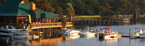 A lakeside scene featuring a colorful boathouse and several boats docked on calm water surrounded by trees.