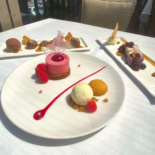 A beautifully plated dessert featuring a pink mousse, ice cream, and garnishes, with additional desserts in the background.