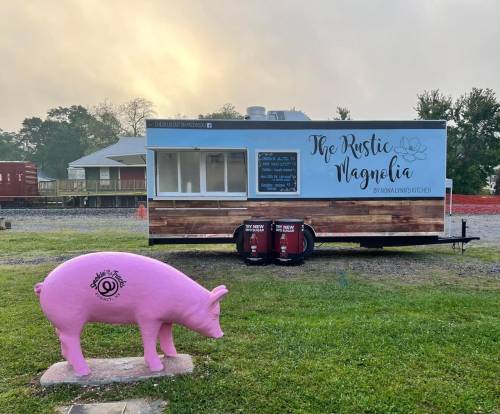 A pink pig statue stands in front of a food truck named "The Rustic Magnolia" in a grassy area.