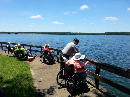 A group of people in wheelchairs and chairs fishing by a lake on a sunny day, with blue skies and green trees in the background.