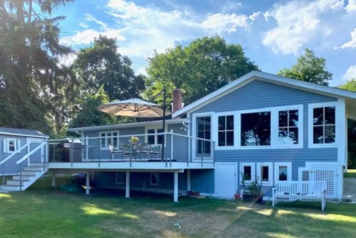 A modern blue house with a large deck, surrounded by greenery and under a bright sky with scattered clouds.