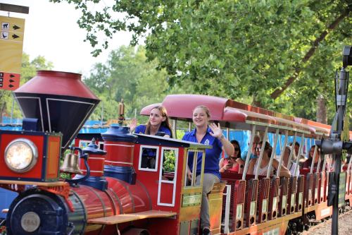 Two women wave from a colorful train as it travels through a park, with trees and a blue sky in the background.