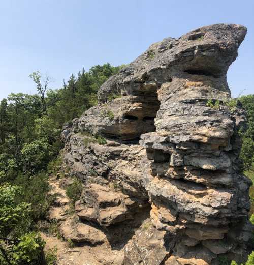 A large, rugged rock formation surrounded by greenery under a clear blue sky.