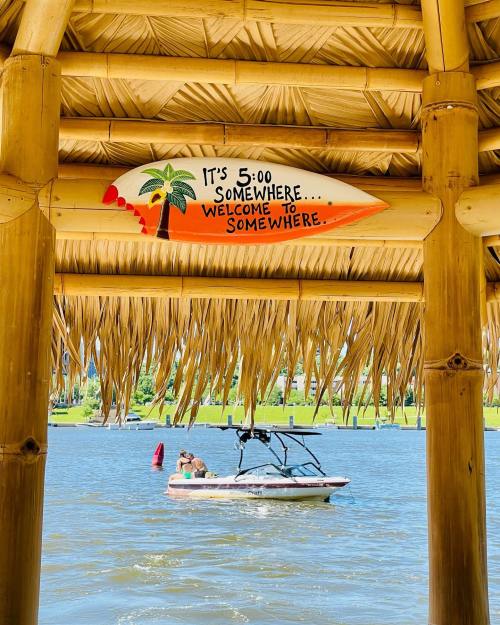 A thatched roof with a sign reading "It's 5:00 Somewhere. Welcome to Somewhere," overlooking a boat on the water.