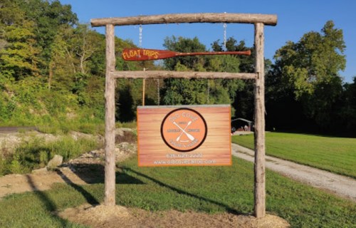 Sign for a float trip company, featuring a wooden frame, paddle, and logo, set in a green outdoor area.