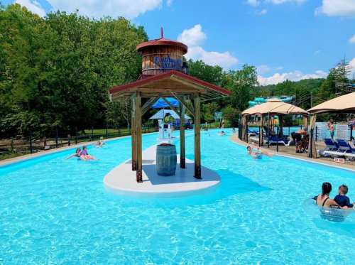 A sunny pool area with a central wooden structure, surrounded by people relaxing in the water and lounging under umbrellas.