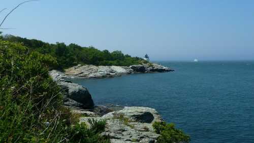 A rocky coastline with lush greenery and a distant lighthouse under a clear blue sky.