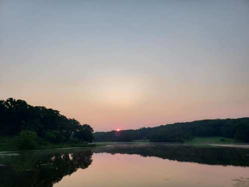 A serene sunset over a calm lake, with trees silhouetted against a pastel sky.