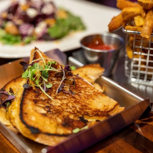 A plate of golden-brown grilled cheese with microgreens, accompanied by a side of fries and dipping sauce.
