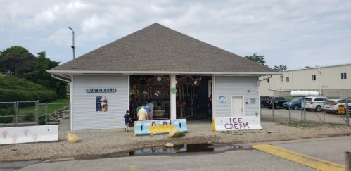 A small ice cream shop with a peaked roof, featuring a sign and people outside on a cloudy day.