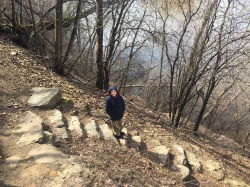 A child stands on a rocky path by a river, surrounded by bare trees and a peaceful landscape.