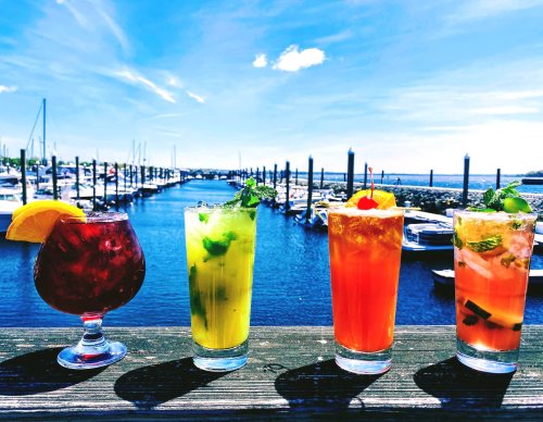 Four colorful cocktails on a railing, with a marina and blue sky in the background.
