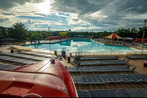 A large, empty swimming pool surrounded by lounge chairs under a cloudy sky, with water slides in the background.
