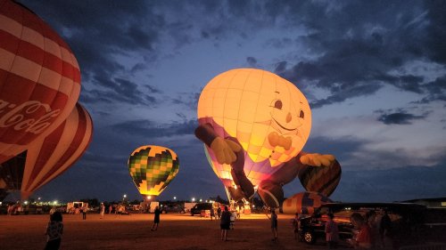 Colorful hot air balloons, including a large smiling character, illuminated against a twilight sky.