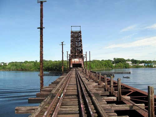 A weathered railway bridge spans a calm river, with tracks leading toward a lift section in the distance.