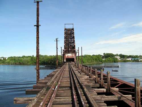 A weathered railway bridge spans a calm river, with tracks leading toward a lift section in the distance.