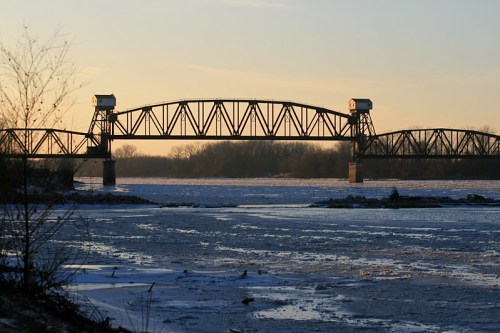 A steel bridge spans a frozen river at sunset, with trees in the background and ice covering the water.