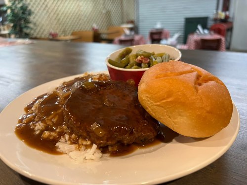 A plate of meatloaf with gravy over rice, served with green beans and a soft roll on the side.