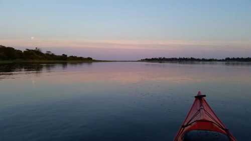 A red kayak on calm water at dusk, with a soft pink sky and a hint of the moon in the background.