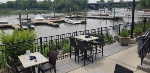 Outdoor dining area by a river, featuring tables, a fence, and boats docked nearby. Lush greenery in the background.