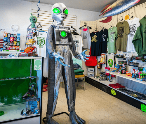 A life-sized alien figure stands in a quirky gift shop filled with alien-themed merchandise and apparel.