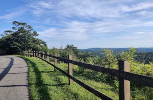 A scenic view of a winding path beside a wooden fence, surrounded by lush greenery and distant hills under a blue sky.