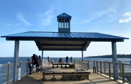 A wooden pier with a gazebo, people enjoying the view of the water under a clear blue sky.