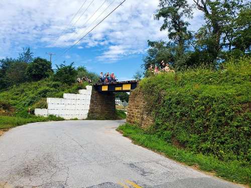 A stone bridge over a road, with people gathered on top, surrounded by greenery and a blue sky.