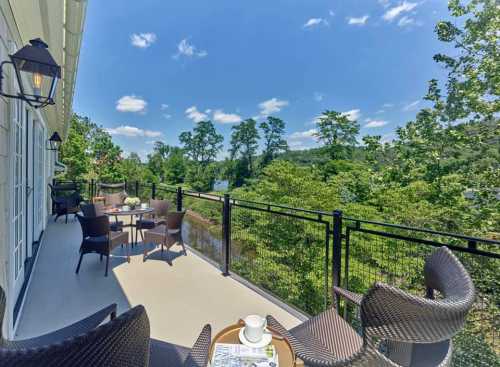 A sunny balcony with chairs and tables overlooking lush greenery and a river under a blue sky.