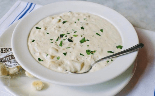 A bowl of creamy soup garnished with parsley, accompanied by a packet of crackers on a white plate.