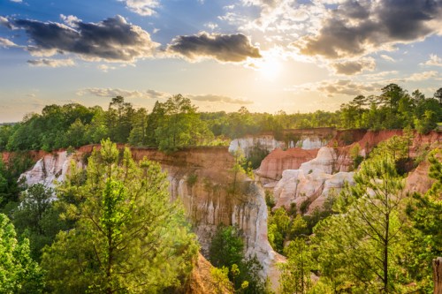 A scenic view of colorful cliffs and lush greenery under a bright sky with clouds at sunset.