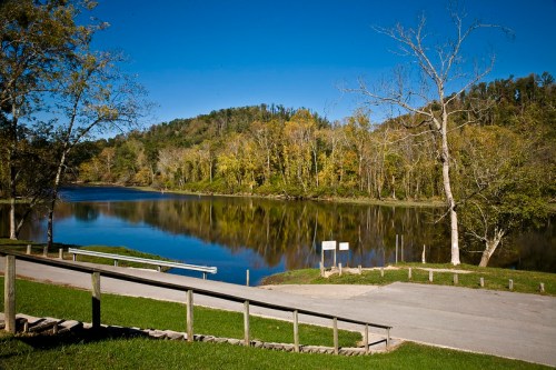 A serene river scene with trees reflecting in the water under a clear blue sky. A grassy area and pathway are visible.