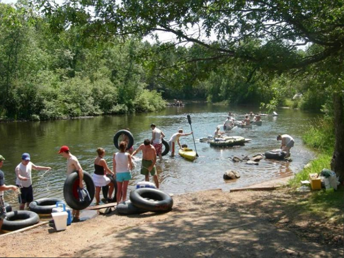 People enjoying a sunny day by a river, preparing to float on inner tubes and kayaks amidst lush greenery.