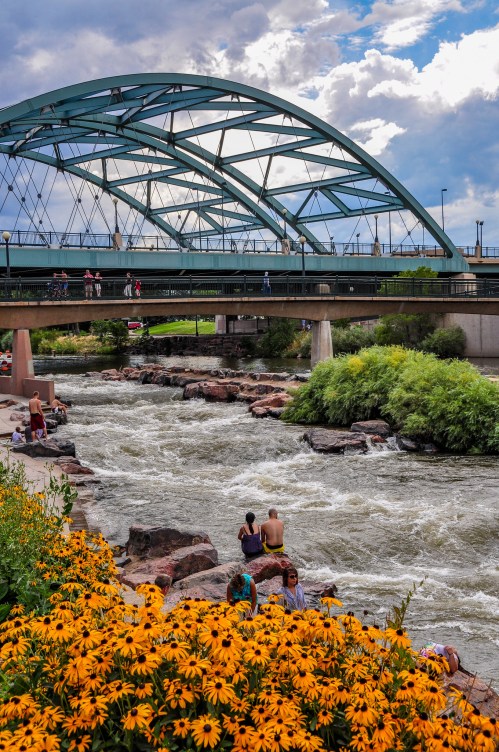 A bridge arches over a river, with people sitting by the water and vibrant yellow flowers in the foreground.